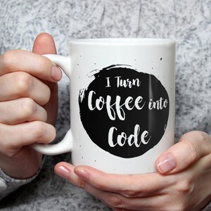 May include: A white ceramic mug featuring a black circular ink-blot design with the text "I Turn Coffee into Code" printed in a white script font. The mug has a smooth handle and a speckled pattern around the central graphic element.