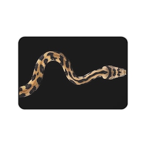 Può includere: Un tappetino rettangolare con una grafica ad alto contrasto di un serpente con un motivo a macchie color marrone chiaro e nero. Il serpente è posizionato in una curva orizzontale a forma di S su uno sfondo nero solido, mostrando squame dettagliate.
