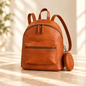 Könnte beinhalten: Ein cognacfarbener Lederrucksack mit einem Hauptreißverschlussfach, einer kleineren Reißverschlusstasche vorne, einem Tragegriff oben und zwei Schultergurten. Ein kleines, rundes, passendes Münztäschchen ist seitlich mit einem Clip befestigt.