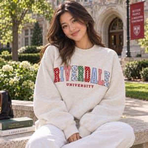 May include: An oatmeal-colored crewneck sweatshirt featuring the word RIVERDALE in large, multi-patterned block letters with red, blue, and green floral, polka dot, and striped prints, with the word UNIVERSITY printed in red below the main text.
