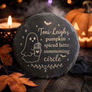 May include: A round, dark grey slate coaster featuring an engraved design of a ghost holding a coffee cup with a heart. The text reads: Toni-Leigh's pumpkin spiced latte summoning circle, surrounded by small stars, bats, and a decorative leaf motif.