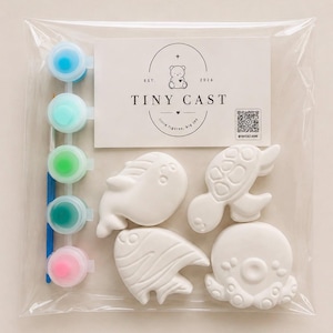 Op de afbeelding: Een knutselset in een doorzichtige plastic zak met vier witte, onbeschilderde zeedierfiguurtjes, een strip met vijf kleine verfpotjes in blauw, turquoise, groen, donker turquoise en roze, en een penseel. Een kaart bevat de tekst TINY CAST en een berenicoon.