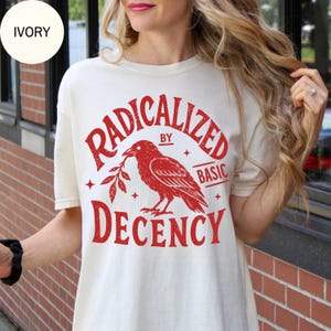 Könnte beinhalten: Ein elfenbeinfarbenes Kurzarm-T-Shirt mit einem roten Grafikdruck eines Raben, der einen Zweig hält. Der Text RADICALIZED BY BASIC DECENCY ist in einer fetten, geschwungenen Schrift gedruckt. Das Design enthält kleine Sternakzente.
