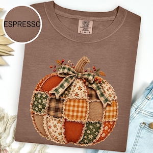 Op de afbeelding: Een espresso-kleurig t-shirt met een afbeelding van een patchwork pompoen. De pompoen is samengesteld uit verschillende stofpatronen, waaronder vichy, bloemen en effen texturen in tinten oranje, groen en bruin, afgewerkt met een groen geruite strik.