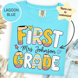 May include: A lagoon blue t-shirt featuring the text FIRST GRADE in large, colorful letters designed to look like pencils and lined notebook paper. The text Mrs. Johnson is printed in the center, accompanied by small heart and pushpin icons.