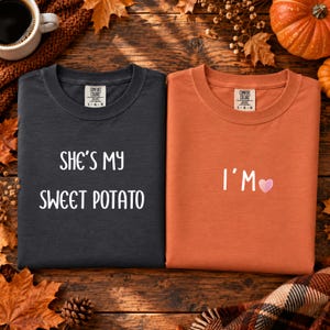Puede incluir: Una camiseta gris oscuro con el texto blanco "SHE'S MY SWEET POTATO" y una camiseta naranja con el texto blanco "I'M" seguido de un pequeño corazón rosa. Ambas camisetas tienen una etiqueta de la marca Comfort Colors en el cuello.