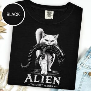 Może przedstawiać: Czarny t-shirt z białą grafiką przedstawiającą kota trzymającego w pysku stworzenie ksenomorfa. Pod grafiką znajduje się napis ALIEN THE SHORT VERSION. Koszulka posiada metkę marki Comfort Colors przy kołnierzu wskazującą rozmiar large.