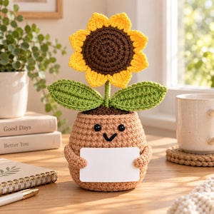 Könnte beinhalten: Eine gehäkelte Sonnenblume in einem beigen Topf mit lächelndem Gesicht, die ein leeres weißes rechteckiges Schild hält. Die Sonnenblume hat eine dunkelbraune Mitte, leuchtend gelbe Blütenblätter und zwei grüne Blätter am Stiel.