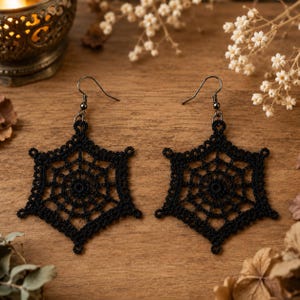 May include: A pair of black earrings featuring a delicate, six-pointed crocheted spiderweb design. Each earring is suspended from a dark metal hook, showcasing intricate thread work that forms a geometric, star-like pattern with a central circular motif.