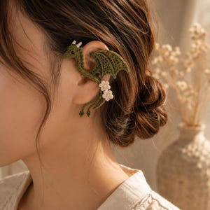 May include: An olive green crocheted ear cuff shaped like a dragon with detailed wings and a long tail. The dragon features small white flower accents with pearl centers along the lower body, wrapping around the outer edge of the ear cartilage.
