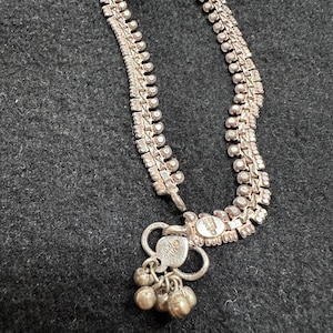 May include: A silver-coloured metal anklet featuring a repeating textured link pattern. The chain terminates in a heart-shaped charm attached to a small ring, which holds three dangling spherical bells. The metal has a slightly oxidised finish.