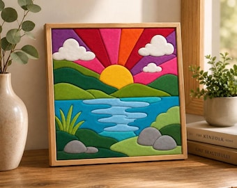 May include: A square wall art piece in a light wood frame featuring a felted landscape scene. The design displays a yellow sun rising over green hills and a blue lake, with white clouds, grey rocks, and vibrant purple and red sunburst rays.