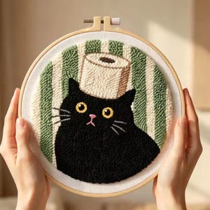May include: A punch needle embroidery hoop featuring a black cat with yellow eyes and a pink nose. The cat has a roll of toilet paper balanced on its head. The background consists of vertical sage green and white stripes on white fabric.