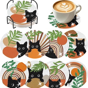 May include: A set of round drink coasters featuring diamond art designs of black cats, green leaves, and orange circles. The coasters are displayed in a black metal holder and individually with a white ceramic coffee cup on top.