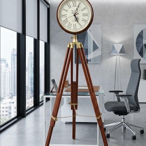 May include: A floor clock featuring a round face with Roman numerals and black hands, mounted on a dark brown wooden tripod stand. The clock head is connected to the legs by brass-colored hardware, and a thin metal chain connects the three legs.