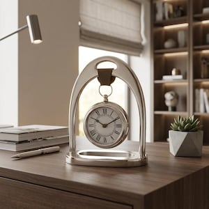 May include: A silver-toned metal desk clock suspended from an arched frame by a brown leather strap. The clock face features a white background with black Roman numerals and two thin black hands, housed in a circular metallic casing.