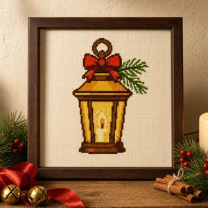 May include: A cross-stitch embroidery in a dark wood frame depicting a yellow lantern with a lit candle inside. The lantern is decorated with a red bow and green pine needles on a cream-colored fabric background with a grid pattern.