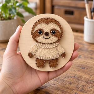 May include: A circular wooden plaque featuring a string art design of a sloth. The sloth is created with brown and cream-colored thread wrapped around small metal nails, depicting a sloth with a smiling face wearing a cream-colored sweater.