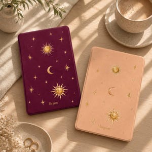 Puede incluir: Dos fundas rectangulares para lector electrónico, una en color burdeos intenso y otra en beige claro, ambas con motivos celestiales dorados de soles, lunas y estrellas. La funda burdeos está personalizada con el nombre Brynn y la beige con Margaret.
