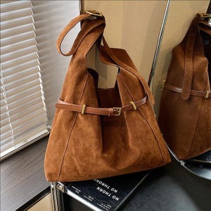 May include: A brown suede hobo bag featuring a slouchy, unstructured silhouette with a single shoulder strap and a thin, adjustable brown leather belt detail across the front, secured with a gold-toned metal buckle and matching hardware accents.