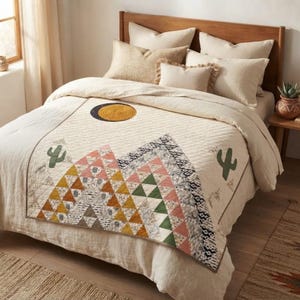 May include: A quilted bedspread featuring a geometric mountain design made of triangles in shades of pink, green, yellow, and white. The quilt includes a circular sun motif and two green cactus icons, paired with several cream-colored pillows.