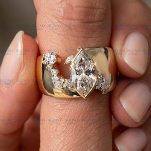 May include: A wide gold band ring featuring a large marquise-cut center stone surrounded by a cluster of smaller round and marquise-cut accent stones. The ring has a polished finish and the text SEGNO is repeated across the image surface.