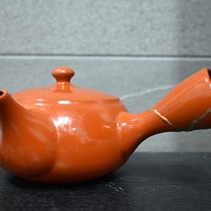 以下が含まれることがあります： 光沢のあるテラコッタ色の陶器製急須で、横手と共蓋が付いています。持ち手の側面には金色の素材で修復された跡が見られ、金継ぎの手法で修復されていることがわかります。
