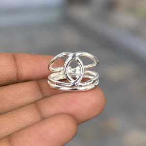 May include: A polished silver ring featuring a multi-band design with interconnected oval loops. The structure consists of several smooth, reflective silver wires that overlap to create a layered, openwork appearance with a continuous metallic finish.