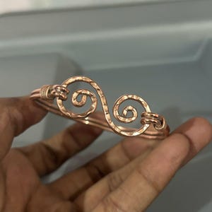 Può includere: Un bracciale rigido in metallo color rame caratterizzato da un design a doppia spirale al centro. La superficie del metallo presenta una texture martellata e le estremità della spirale sono fissate con sottili avvolgimenti di filo su entrambi i lati.
