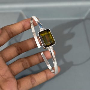 May include: A silver cuff bracelet featuring a large, rectangular, faceted olive-green gemstone set in a polished metal frame. The band is smooth and open-ended, connecting to the central stone setting with delicate wire-wrapped silver detailing.