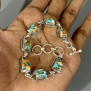 Könnte beinhalten: Ein silbernes Kettenarmband mit sechs rechteckigen Gliedern, die jeweils mit einem Mosaik aus türkisfarbenen, orangefarbenen und weißen Steinfragmenten eingelegt sind. Das Armband verfügt über einen Knebelverschluss und Einstellringe.