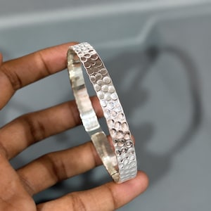 May include: A silver-colored bangle bracelet featuring a textured surface with a repeating pattern of small, circular indentations. The metal has a polished, reflective finish that catches the light across its curved, rigid band structure.