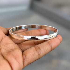 May include: A polished silver-tone bangle bracelet with a smooth, rounded surface and a continuous circular design. The metal has a high-shine, reflective finish that catches the light, creating a clean and simple aesthetic for this wrist accessory.