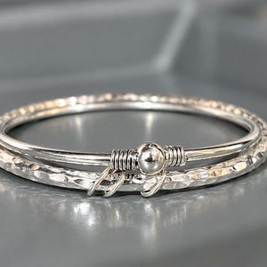 May include: A silver bangle bracelet featuring a smooth inner band and a textured, hammered outer band. The two bands are connected by a central silver bead with wire-wrapped detailing and three small, dangling silver rings attached to the side.