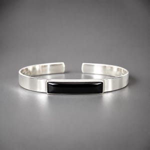 May include: A silver cuff bracelet featuring a smooth, polished finish and an open-ended design. The center of the bracelet is inlaid with a long, rectangular, glossy black stone that sits flush within the metallic band, creating a sleek contrast.