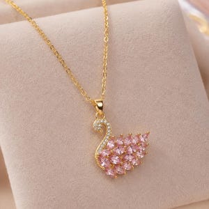 Può includere: Una collana color oro con un ciondolo a forma di cigno. Il collo del cigno è incastonato con piccole pietre chiare, mentre il corpo è composto da molteplici pietre rosa taglio marquise disposte a formare le piume su una montatura dorata.