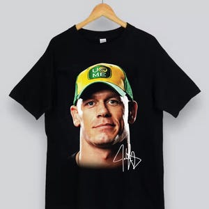 Puede incluir: Una camiseta negra de manga corta con un estampado gráfico de un hombre que lleva una gorra de béisbol amarilla y verde con el texto U CAN'T SEE ME. Debajo del retrato hay una firma blanca estilizada. La prenda cuelga de una percha de madera.
