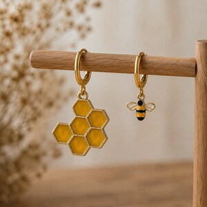Puede incluir: Un par de pendientes de aro dorados con dijes desiguales. Un pendiente tiene un colgante de panal de esmalte amarillo, mientras que el otro presenta un pequeño dije de abeja con detalles de esmalte negro, amarillo y blanco en alas y cuerpo.