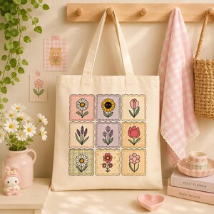 May include: A cream-colored canvas tote bag featuring a grid of nine square patches, each displaying a different colorful flower illustration. The flowers include daisies, sunflowers, and tulips in various pastel shades, arranged in a three-by-three pattern.