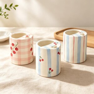 May include: Three cylindrical ceramic toothbrush holders with open tops and a small circular hole. One features a pink gingham pattern with red cherries, while the other two display light blue vertical stripes accented with red cherry motifs.