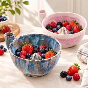 Può includere: Due ciotole spremiagrumi in ceramica, una con smalto blu e marrone screziato e l'altra con smalto rosa e bianco, ciascuna dotata di un cono centrale e di un manico laterale, riempite con fragole, lamponi e mirtilli freschi.