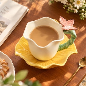 May include: A ceramic cup and saucer set featuring a cream-coloured cup with a wavy rim and a yellow leaf-shaped saucer. The cup handle is sculpted as a green vine topped with a pink butterfly. The cup is filled with a light brown liquid beverage.