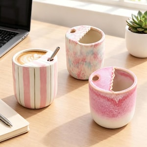 Puede incluir: Tres tazas cilíndricas de cerámica con un borde dentado y un pequeño orificio circular para una pajita. Una taza presenta rayas verticales rosas, otra un acabado jaspeado en rosa y azul, y la tercera un degradado de color rosa.