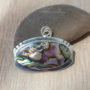 Könnte beinhalten: Ein ovaler Anhänger mit einer Einlage aus poliertem Abalone-Muschelgehäuse in irisierenden Violett-, Grün- und Blautönen. Die Muschel ist in einen silberfarbenen Metallrahmen mit glattem, geschwungenem Rand und einer kleinen Öse oben gefasst.