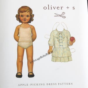 May include: An Oliver + S sewing pattern envelope for an Apple-Picking Dress. The white cover features an illustration of a child in undergarments and a separate illustration of a dress with a bow, buttons, and an apple. Sizes 6-12M to 4.