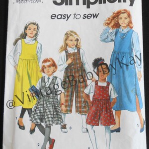 May include: A Simplicity 7953 sewing pattern envelope for sizes AA (7-10) featuring illustrations of four outfits: a yellow jumper, a plaid dress, plaid overalls, and a red floral romper, all worn over white shirts with various footwear.