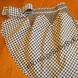 May include: A black and white gingham fabric apron featuring a smocked waistband and a side tie closure. The fabric is printed with the text @VintageBabyByKay across the front, displaying a classic checkered pattern throughout the garment.