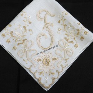 May include: A white square handkerchief featuring intricate gold-toned embroidery of a large letter F in the center, surrounded by floral vines, bows, and a scalloped edge. The text @VintageBabyByKay is embroidered in the center of the fabric.