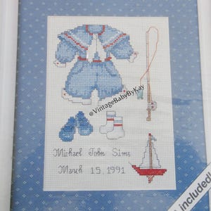 May include: Framed cross-stitch artwork featuring a baby's outfit, shoes, and a sailboat, all in blue and white. Includes a fishing rod and the name "Michael John Sims" and date "March 15, 1991". The artwork is framed in a white frame.