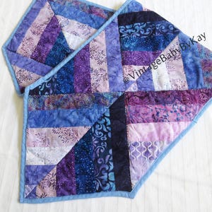 May include: Two quilted table runners in shades of blue and purple. The runners are made of fabric strips sewn together in a geometric pattern. The edges are finished with a light blue border. The text "VintageBabyByKay" is visible.